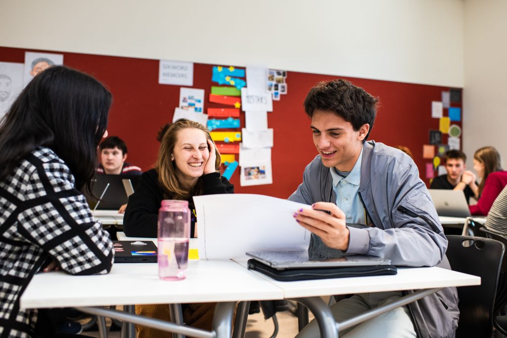A male teacher at a classroom table tacking to two female students, and discussing some work on a sheet of peper.